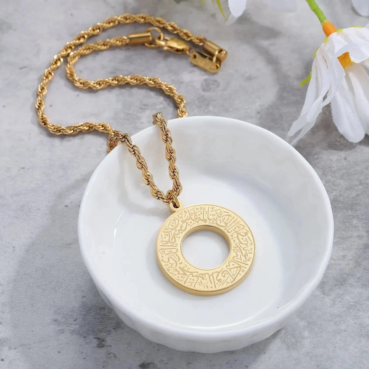 Gold-plated circular pendant necklace with Arabic calligraphy in a white ceramic bowl.