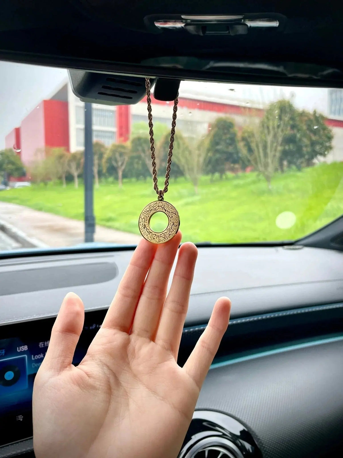 A gold circular pendant necklace hanging from a car rearview mirror.